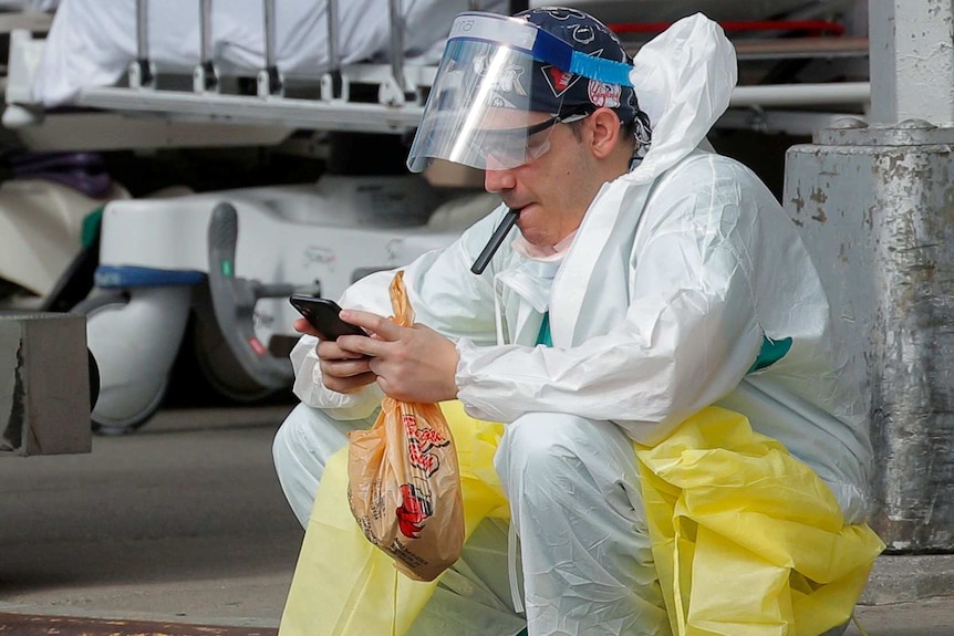 A man in hospital clothes and a protective mask sits on the roadside looking at his phone