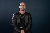 A man with a bald head and beard wears a black jacket and sits against a black background