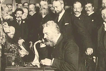 A black and white photo of Bell on the telephone in New York (calling Chicago) with an audience in the background.