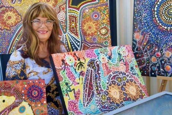 A blonde woman in front of Indigenous art smiles while holding up another painting