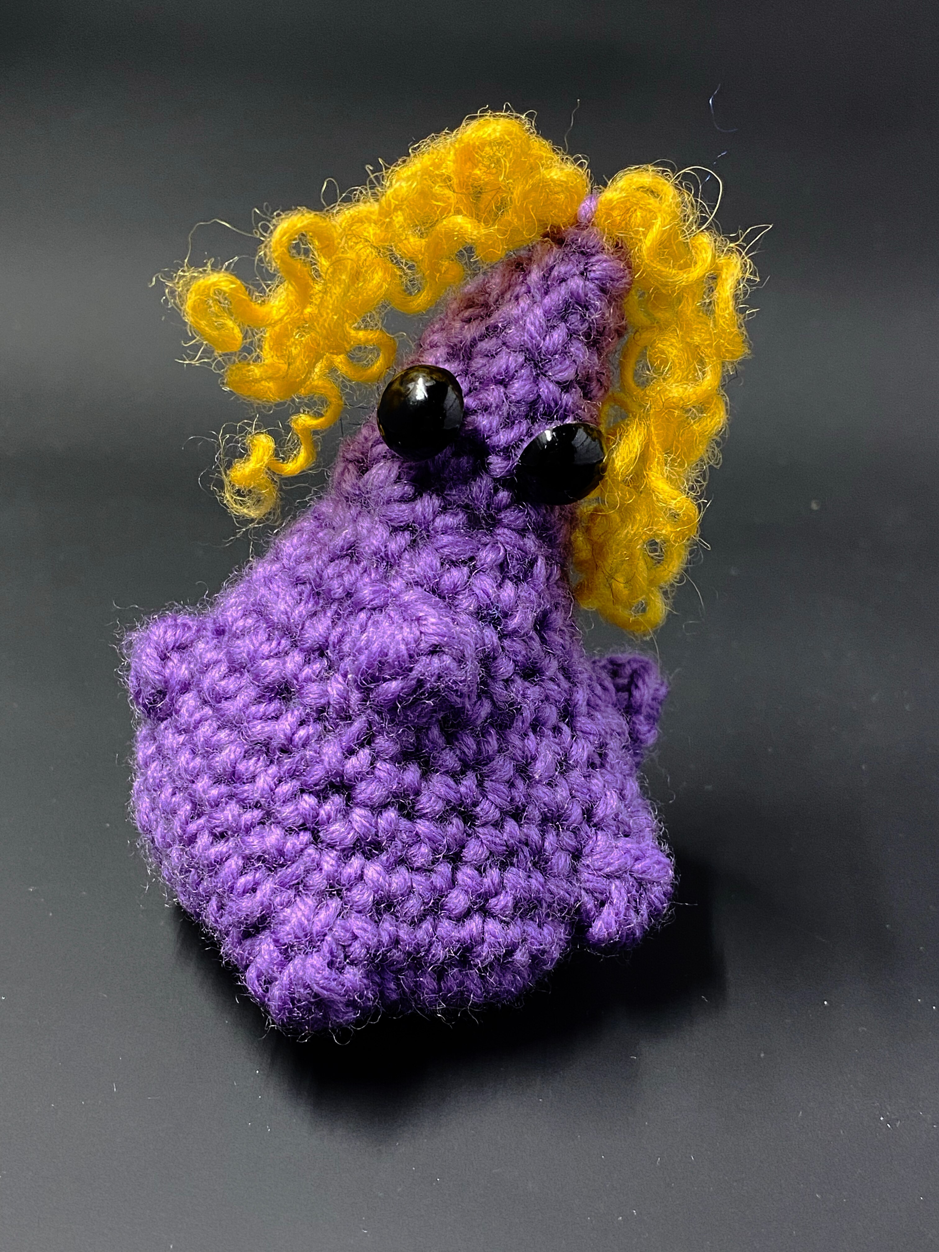Picture of a crocheted organ, which is a purple oblong shape of a tumour with black button eyes and curly yellow knitted hair