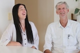 Young woman, long black hair, older man, grey hair and beard, seated at a kitchen table and laughing.