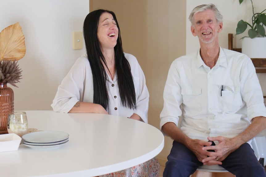 Young woman, long black hair, older man, grey hair and beard, seated at a kitchen table and laughing.