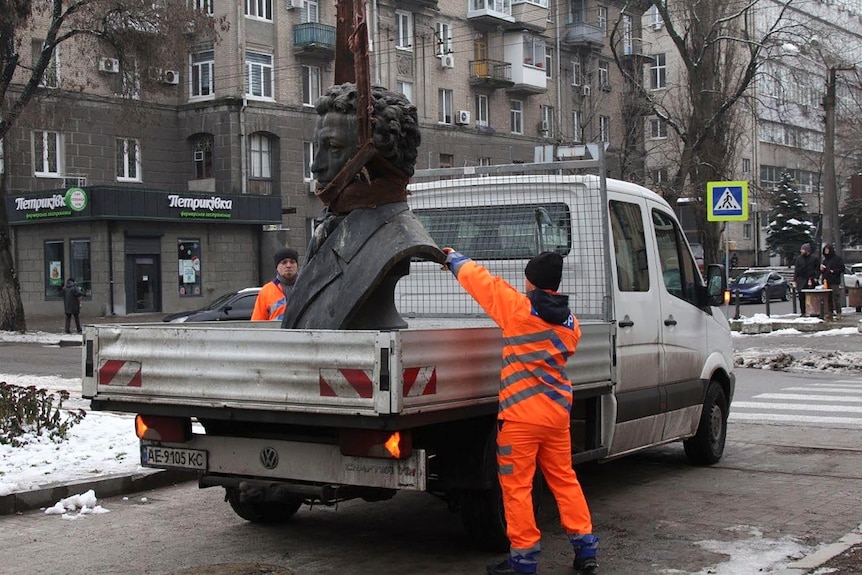 Two men in orange hi-vis clothing assist a large metal bust of a distinguished-looking man on to the back of a ute.