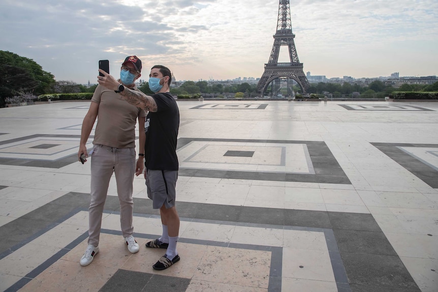 Two men in blue face masks take a selfie near the Eiffel Tower.