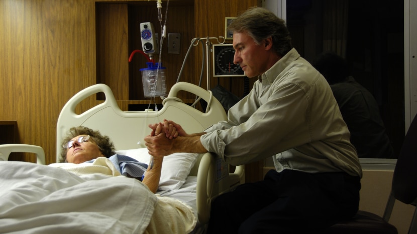 Man at bedside holding of elderly woman in hospital bed