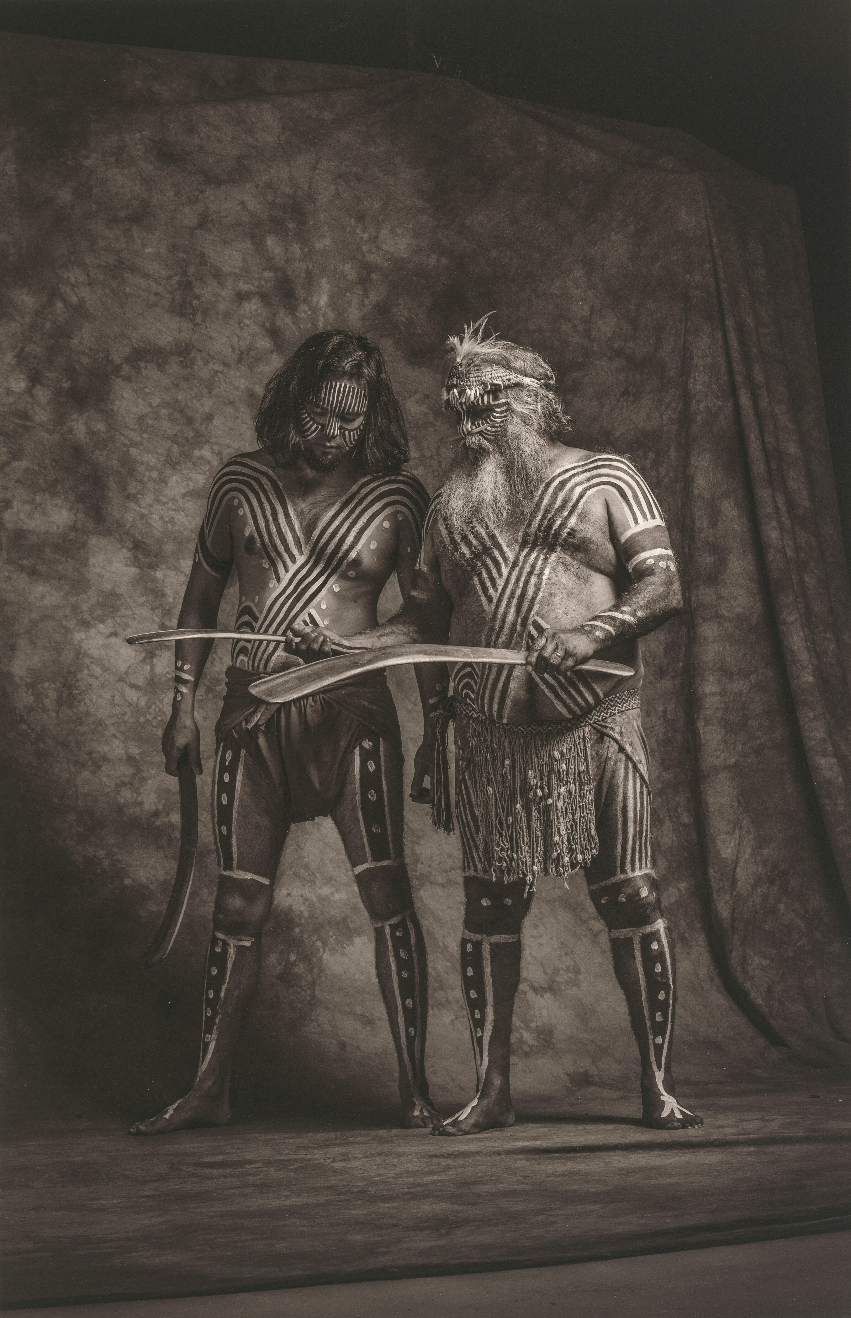 Sepia-tone photograph of young and older Ngarrindjeri men painted-up and dressed for ceremony, against a dropcloth.