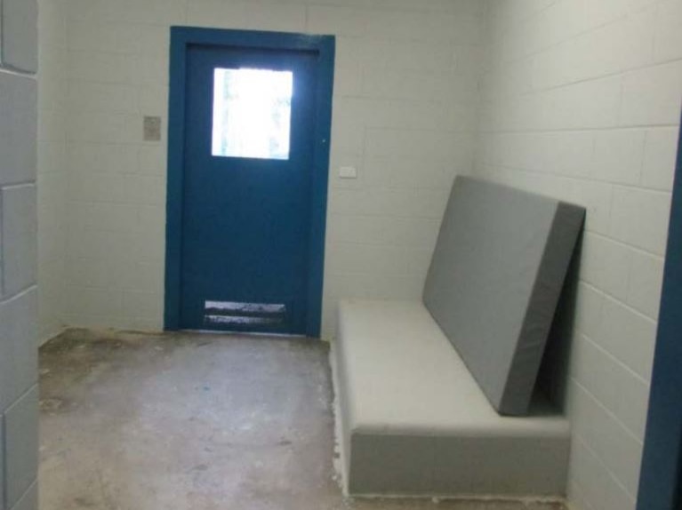 Empty cell with a blue door and grey concrete bed and mattress.