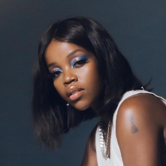 Tkay Maidza infront of a navy background wearing a white tank top, a silver chain and blue eyeshaddow