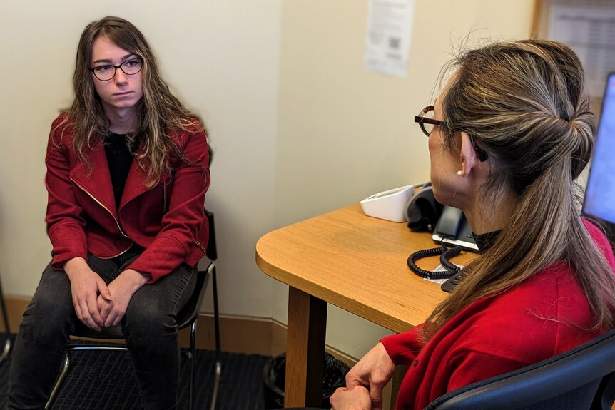 A young woman with dark hair sits in a doctor's office looking seriously at a woman in glasses.
