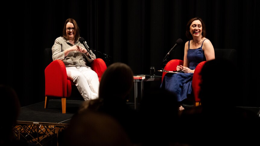 Two women sit on red chairs on a dim stage, smiling.