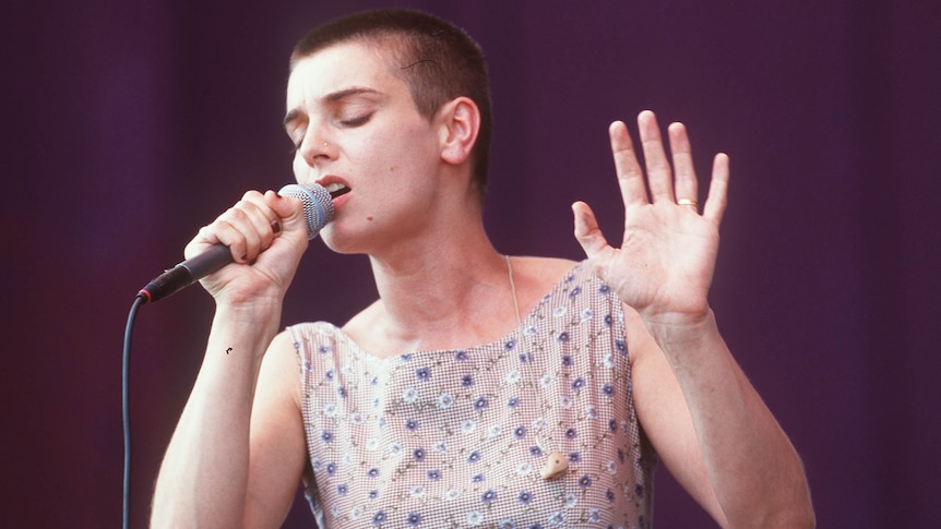 Woman with shaved head in floral dress sings into microphone 