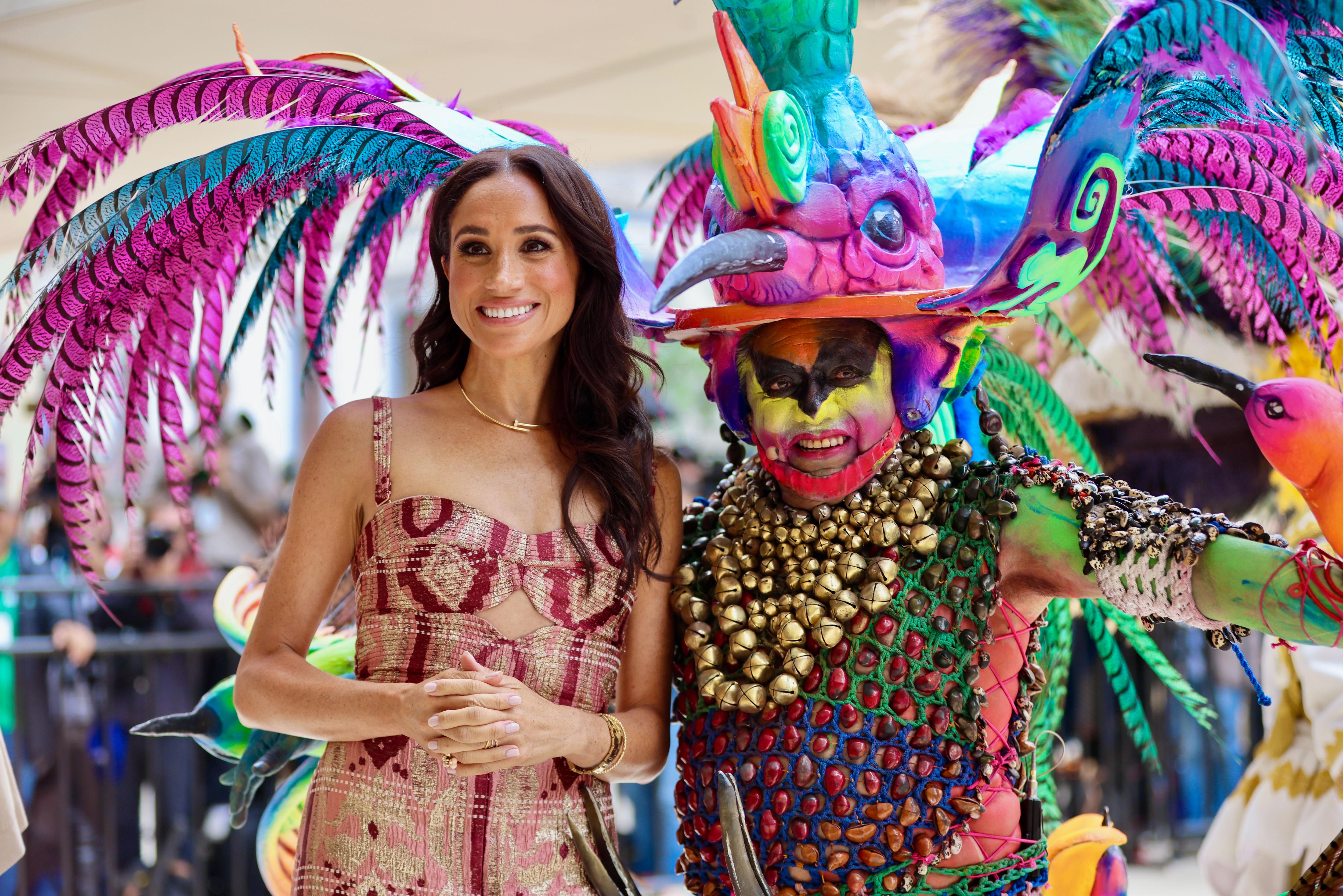 Meghan smiles as she stands next to a performer dressed as a giant, colourful bird.