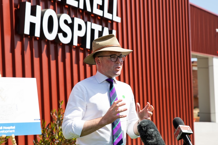 A man with glasses and a hat stands in front of microphones outside a red building with Hospital written on it.