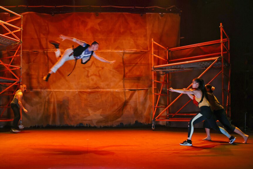 On a stage, a man in a harness swoops through the air. Two Asian women lunge forward towards him. An Arab man cowers.