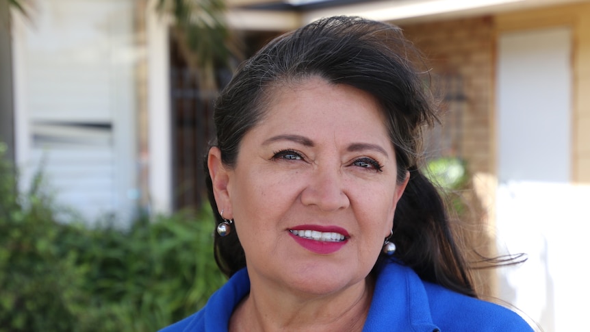 A woman stands in front of a house, smiling at the camera wearing a blue shirt 