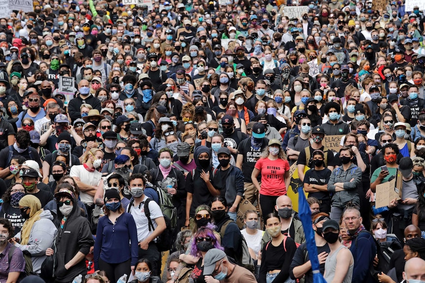 A crowd of thousands of people, many wearing face masks, stand together filling a wide city street.