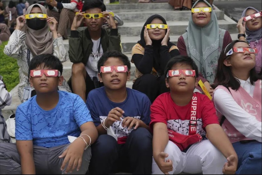 Young Indonesian people sit on a set of steps, all wearing special eyewear to view a solar eclipse.