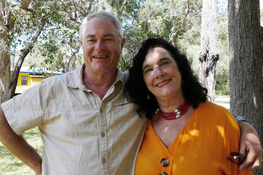 A man and woman stand together with bushland and a yellow cottage in the background