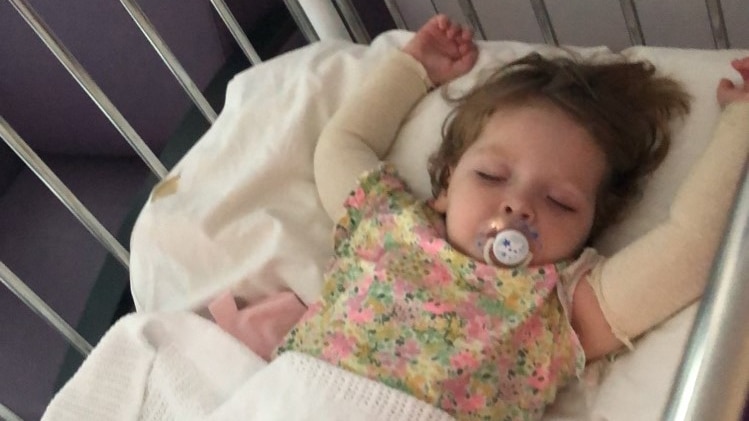 a young girl lays in a hospital bed with bandaged arms