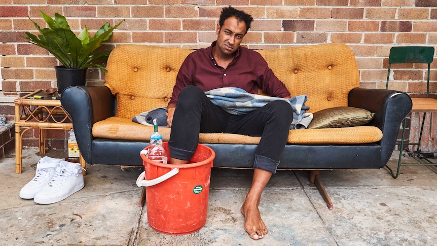 A man wearing a maroon shirt and rolled up jeans sitting on a couch with his foot in a bucket.