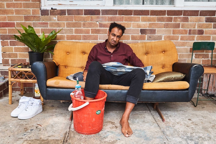 A man wearing a maroon shirt and rolled up jeans sitting on a couch with his foot in a bucket.