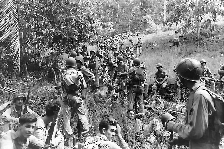 Troops lie in the grass in the side of a hill in Guadalcanal.