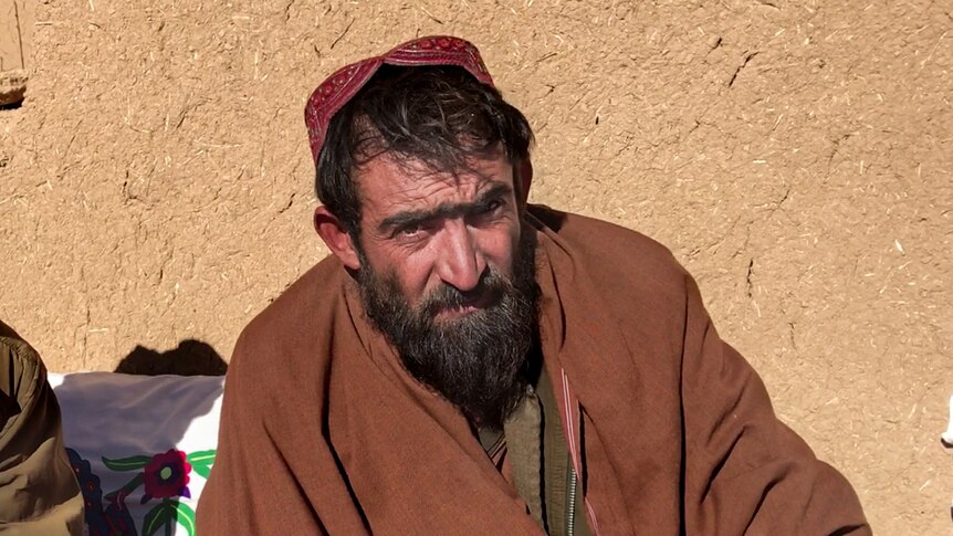 Sayed Jan, beared with black hair wearing a red cap, sits in front of a light brown wall outside.