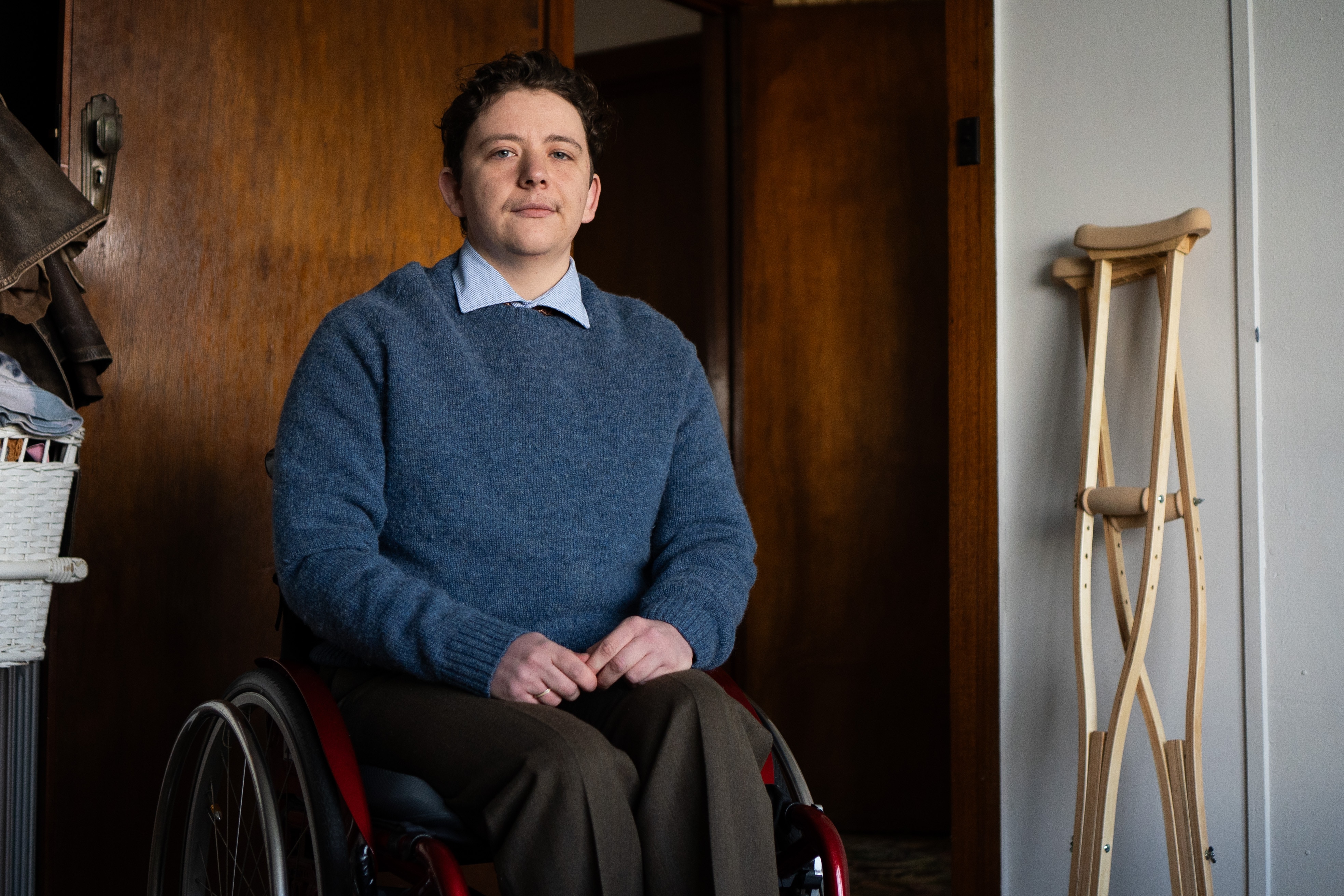 A young man sitting in a wheelchair. Crutches lean against the wall.