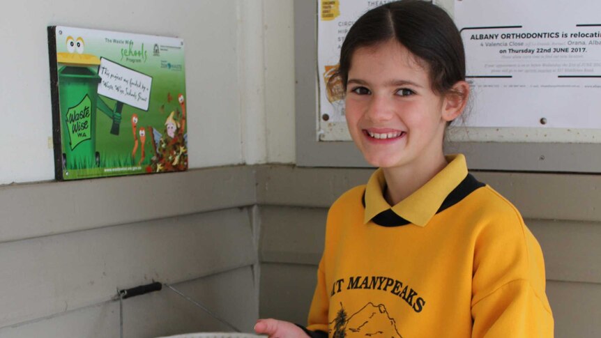 A young girl in school uniform holds batteries in her hands next to a bucket