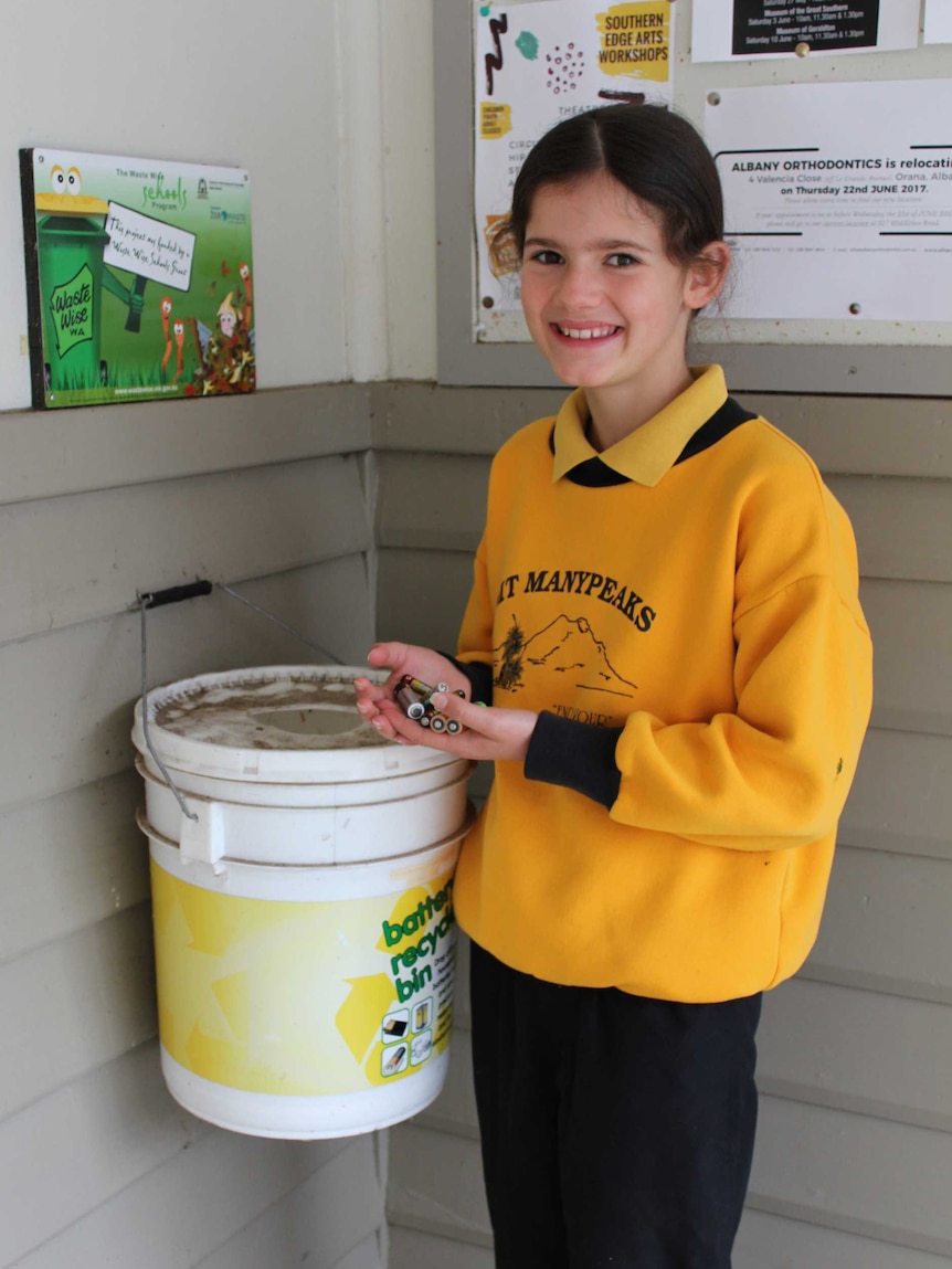 A young girl in school uniform holds batteries in her hands next to a bucket