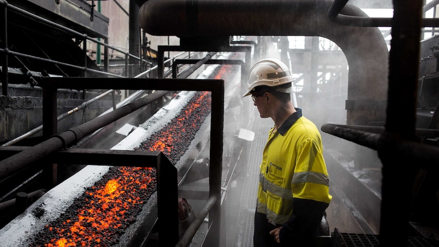A man wearing safety equipment looks at a conveyer belt of molten ore.