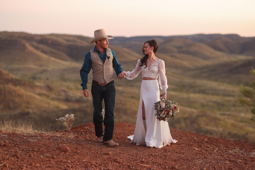 A bride in a white dress with long brown hair holds her grooms hand in an outback landscape.  He wears blue shirt and cream vest