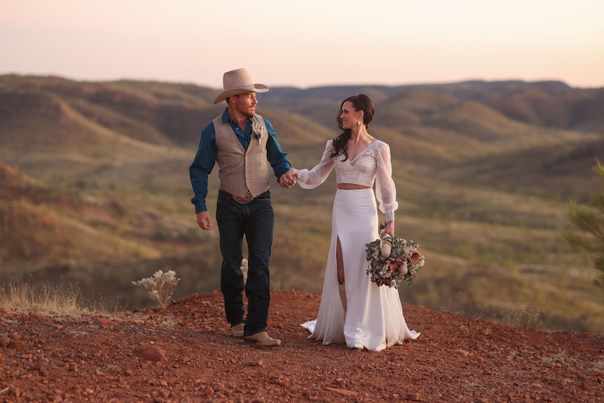 A bride in a white dress with long brown hair holds her grooms hand in an outback landscape. He wears blue shirt and cream vest