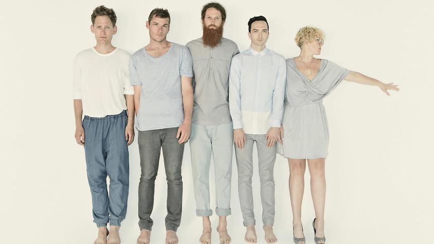 The five members of Architecture In Helsinki standing in a line, wearing blue and grey linen. Kelly has her arm outstretched