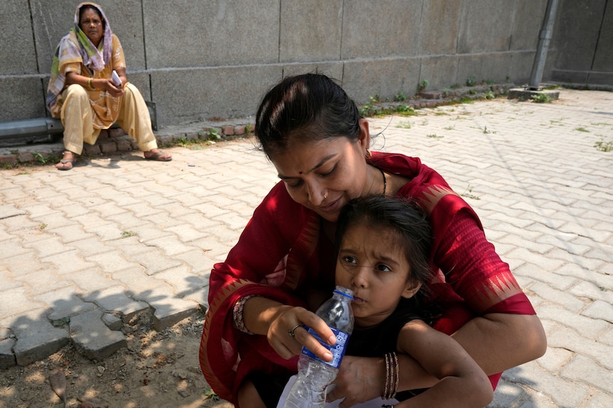 A middle-aged woman leans down to give a water bottle to her daughter in a brightly lit courtyard.