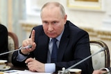 Vladimir Putin points his right ring finger upwards as he sits at a table near a mic.