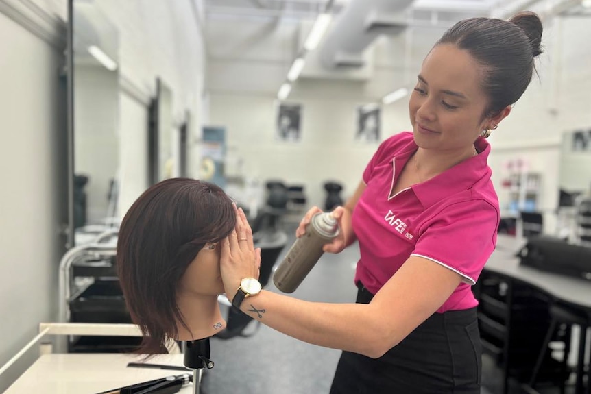 A woman in a pink polo shirt styles a mannequins hair. 