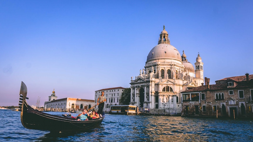 View of imposing cathedral with cupola across the choppy water of a Venice canal. Gondola with tourists in the foreground.