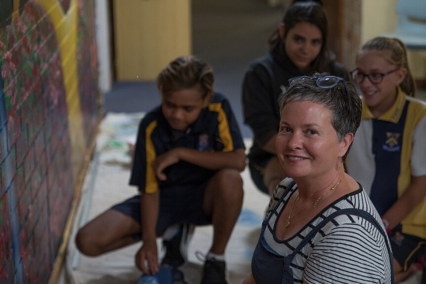 A woman sitting down near a painted wall with three school children sitting down beside her