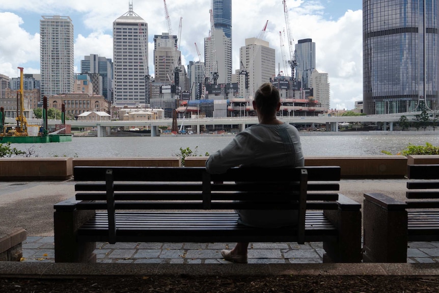 An unidentifiable woman sits on  park bench looking out over Brisbane's CBD.