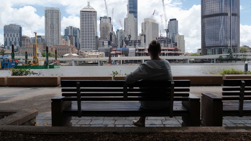 An unidentifiable woman sits on  park bench looking out over Brisbane's CBD.