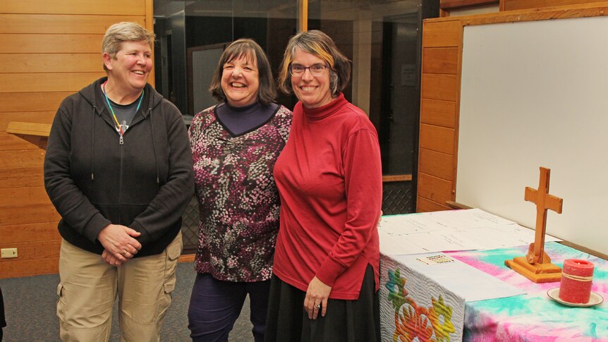 Three women smiling next to a colourful table with a cross on it.