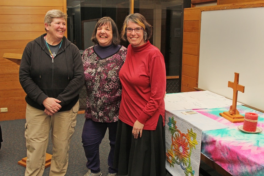 Three women smiling next to a colourful table with a cross on it.