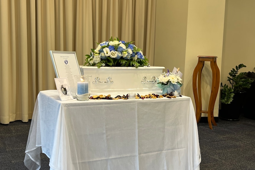A small white coffin topped with flowers sits on a table at a funeral.