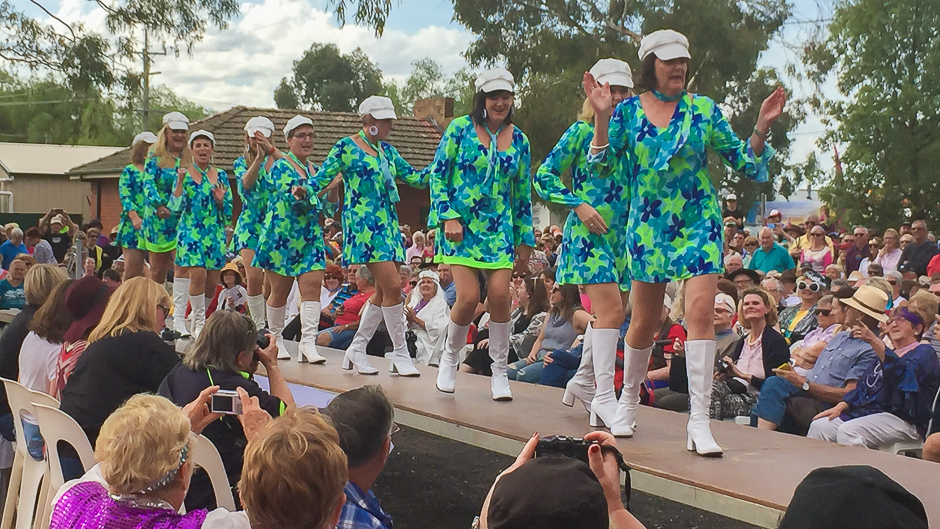 A line of women in the same blue and green mini dresses and white boots walking down a stage