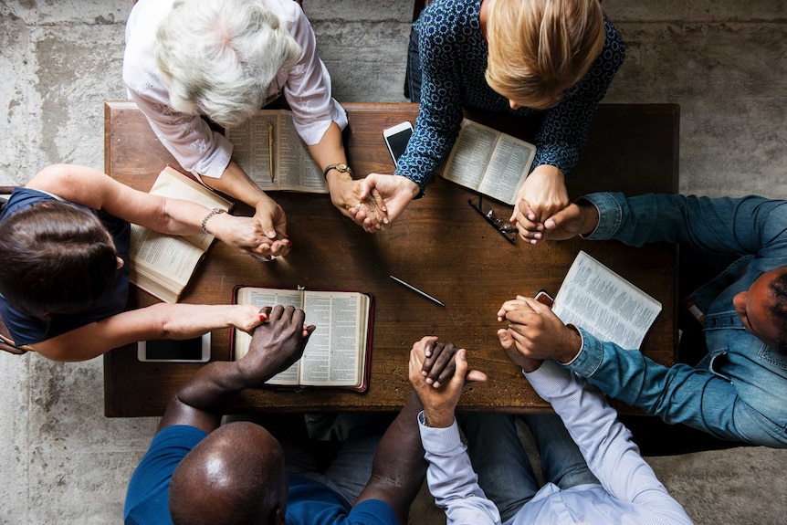 A diverse group of people holds hands over bibles at a table.