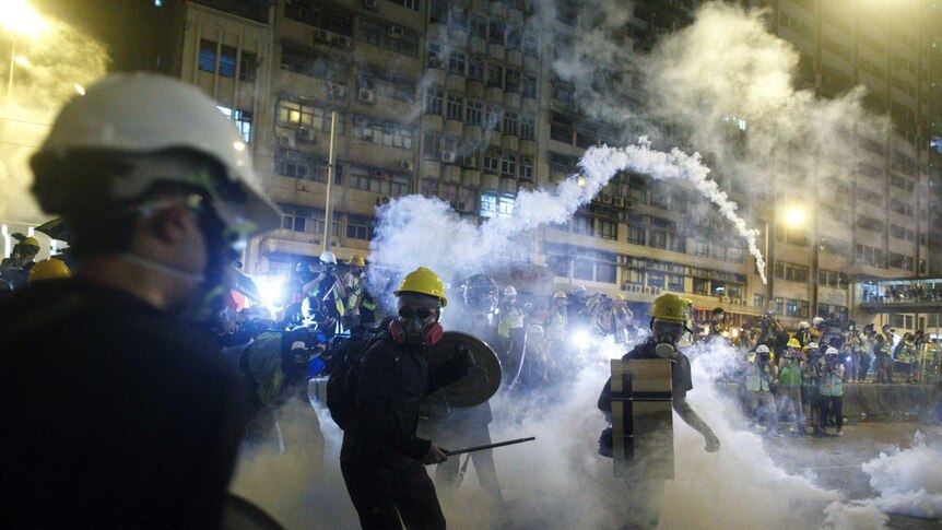 Viewed behind the shoulders of protestors wearing gas masks and hard hats, you look down a street which is filled with tear gas.