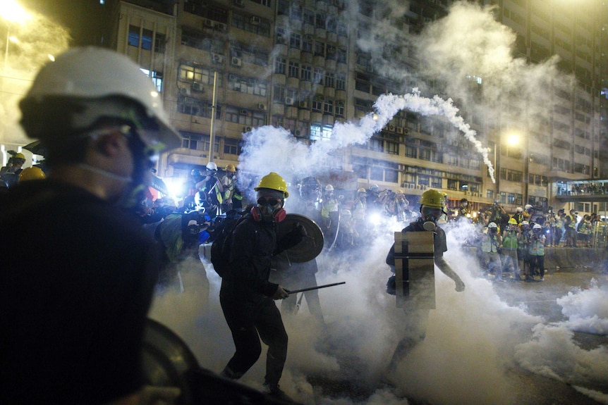 Viewed behind the shoulders of protestors wearing gas masks and hard hats, you look down a street which is filled with tear gas.