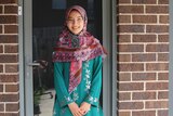A young woman in a bright pink patterned hijab stands outside the front door of her brown brick home.